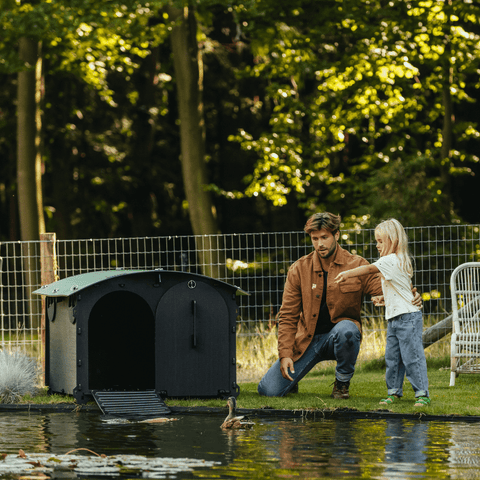 Sfeerfoto Nestera kunststof groot konijnenhok groot eendenhok vooraanzicht, aan het water op het gras met eenden en een vader en dochter.