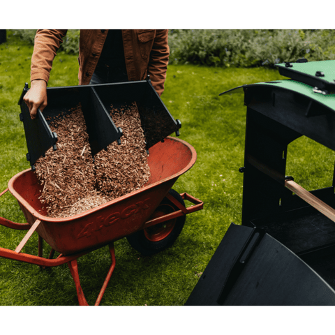 Sfeerfoto van man die zijcompartiment van Nestera kunststof kippenhok groot op poten leegt in een kruiwagen, op het gras in een achtertuin. Van het kippenhop rechts op de foto is de achterzijde verwijderd.