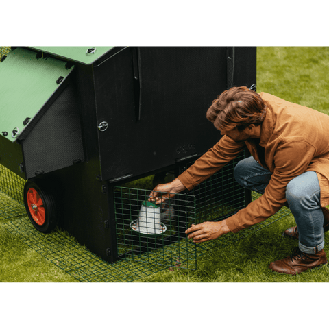 Sfeerfoto Nestera kunststof kippenhok groot met 2 meter ren, schuin achteraanzicht in een achtertuin op het gras. Een man opent de ren en plaatst een voederbak in de ren onder het kippenhok.