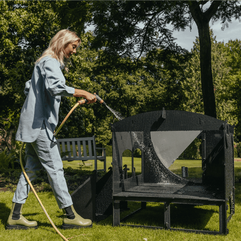 Sfeerfoto Nestera kunststof kippenhok groot op poten vooraanzicht, in een achtertuin op het gras. De achterzijde van het kippenhok is verwijderd en er staat een vrouw naast het kippenhok die het kippenhok schoonspuit met een tuinslang.
