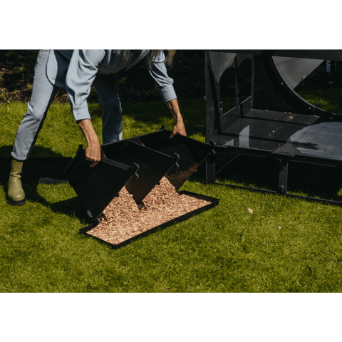 Sfeerfoto Nestera kunststof kippenhok groot op poten schuin vooraanzicht, in een achtertuin op het gras. De achterzijde van het kippenhok is verwijderd en er staat een vrouw naast het kippenhok die het zijcompartiment vol met zaagsel leegt in een platte bak.